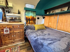 Une chambre à coucher avec un lit, une commode en bois, des étagères remplies de bouteilles, une tapisserie et une fenêtre avec des rideaux.