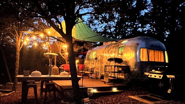 Un camping-car à l’arrêt, doté d’un espace repas couvert, d’une table et de chaises, le tout entouré d’arbres et illuminé de guirlandes lumineuses.