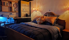 Una camera da letto con un letto, una scrivania con una sedia, una libreria, una sveglia e una lampada a muro.