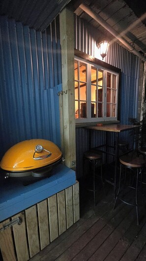 Interior rústico de una cabaña con un banco de madera, una estufa a gas y una ventana con vistas a una terraza de madera.