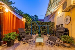 Un espacio al aire libre con sillas y una mesa, rodeado de plantas en macetas y una estructura metálica azul.