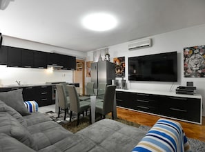 Un saló modern amb un sofà, una taula de dinar amb cadires, un televisor de pantalla plana i una cuina amb armaris i aire condicionat.