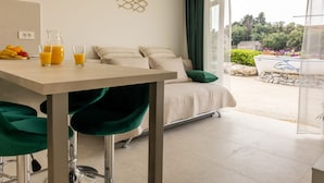 A modern hotel room with a bed, a dining table with breakfast items, a green chair, and a view of the outdoors. - Apartman Helena Korčula (Blato)