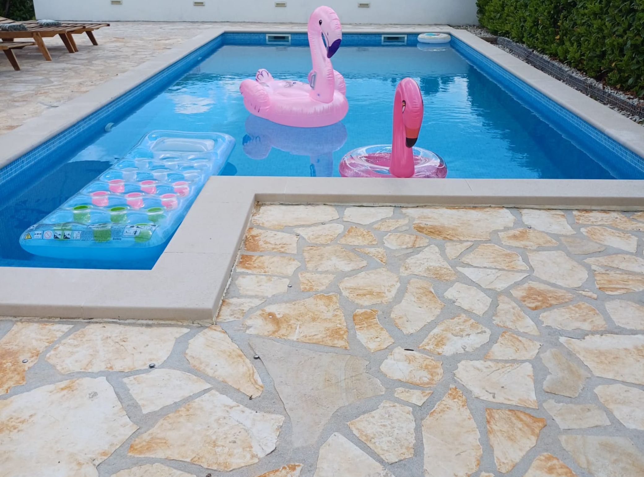A swimming pool with pink flamingo floats, a floating pool table, and stone tiles.