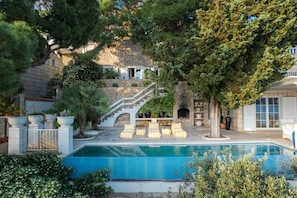 A pool area with lounge chairs, a stone staircase, and a view of a building with a stone facade. - Villa Mey's Place (Dubrovnik)