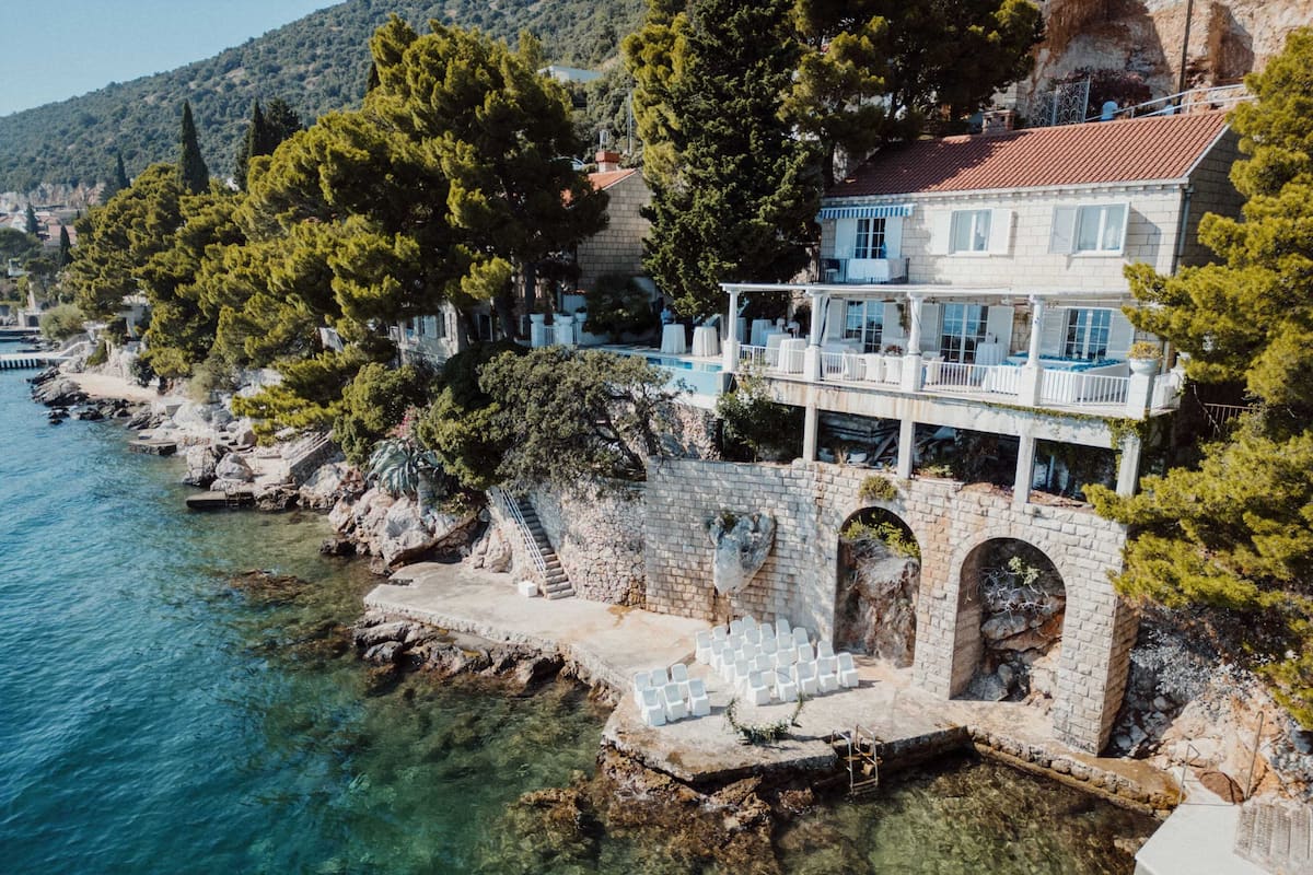 A coastal hotel with a stone staircase leading to a terrace, clear blue sea, and lush greenery.