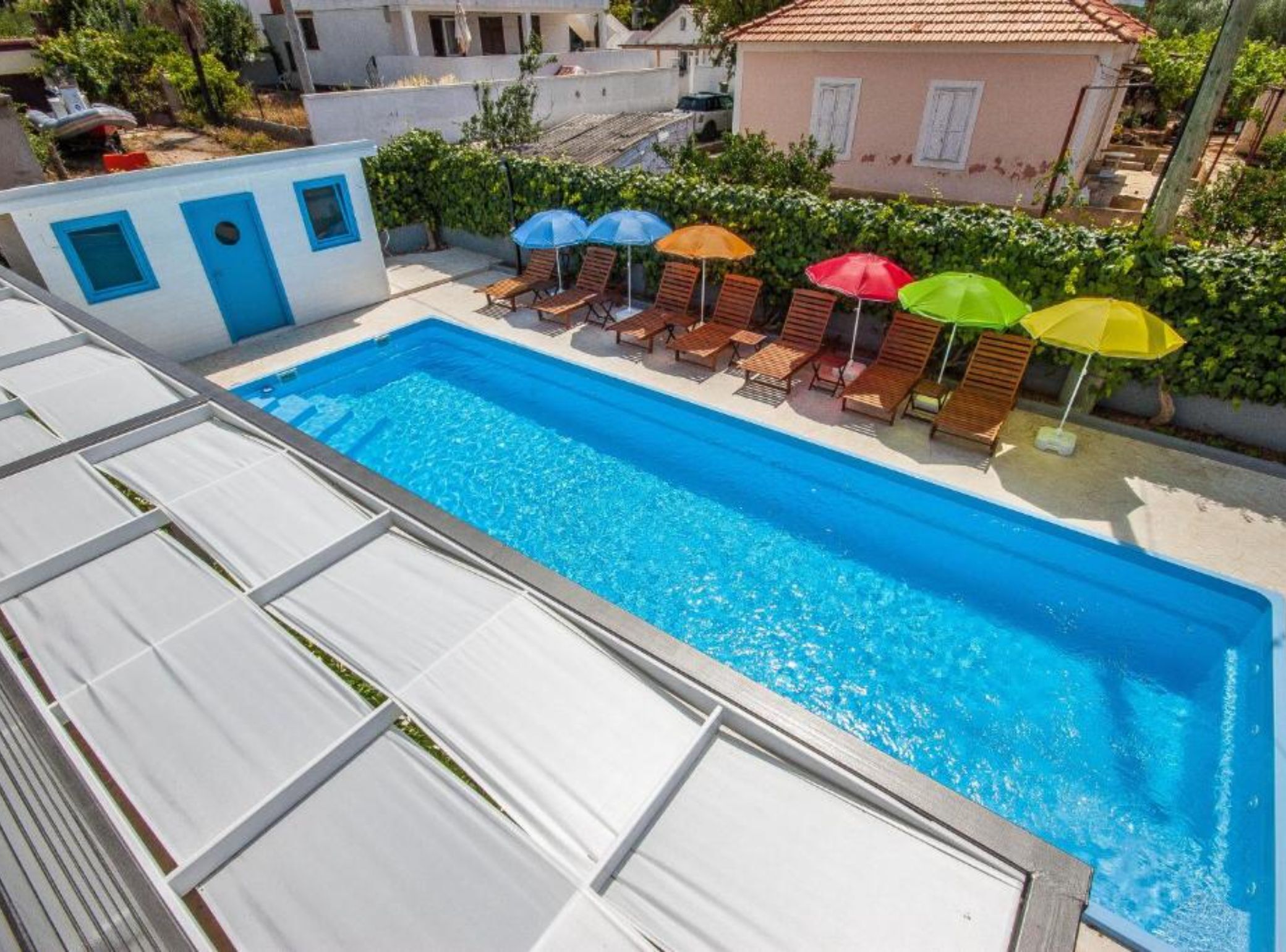 Ein Schwimmbecken mit Liegestühlen und Sonnenschirmen, umgeben von Grünflächen und Gebäuden.