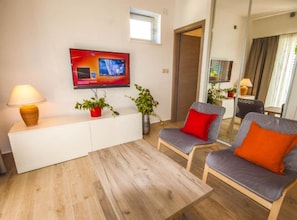 Ein modernes Wohnzimmer mit einem Flachbildfernseher, zwei Sesseln mit roten Kissen, einem weißen Konsolentisch, einer Stehlampe und einem Topf mit Pflanze.