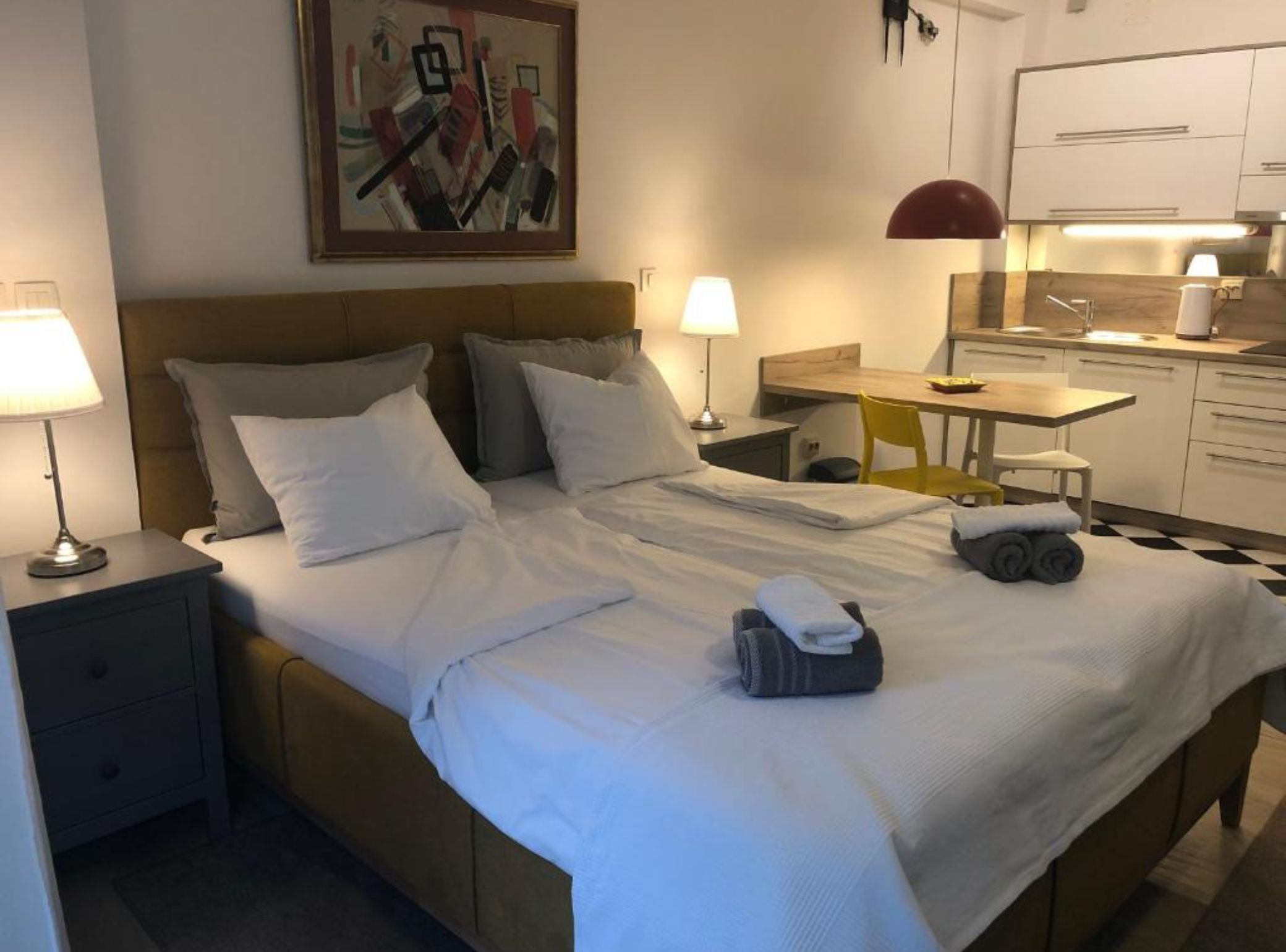 Habitación de hotel con cama, lámparas de noche, escritorio, silla y zona de cocina.