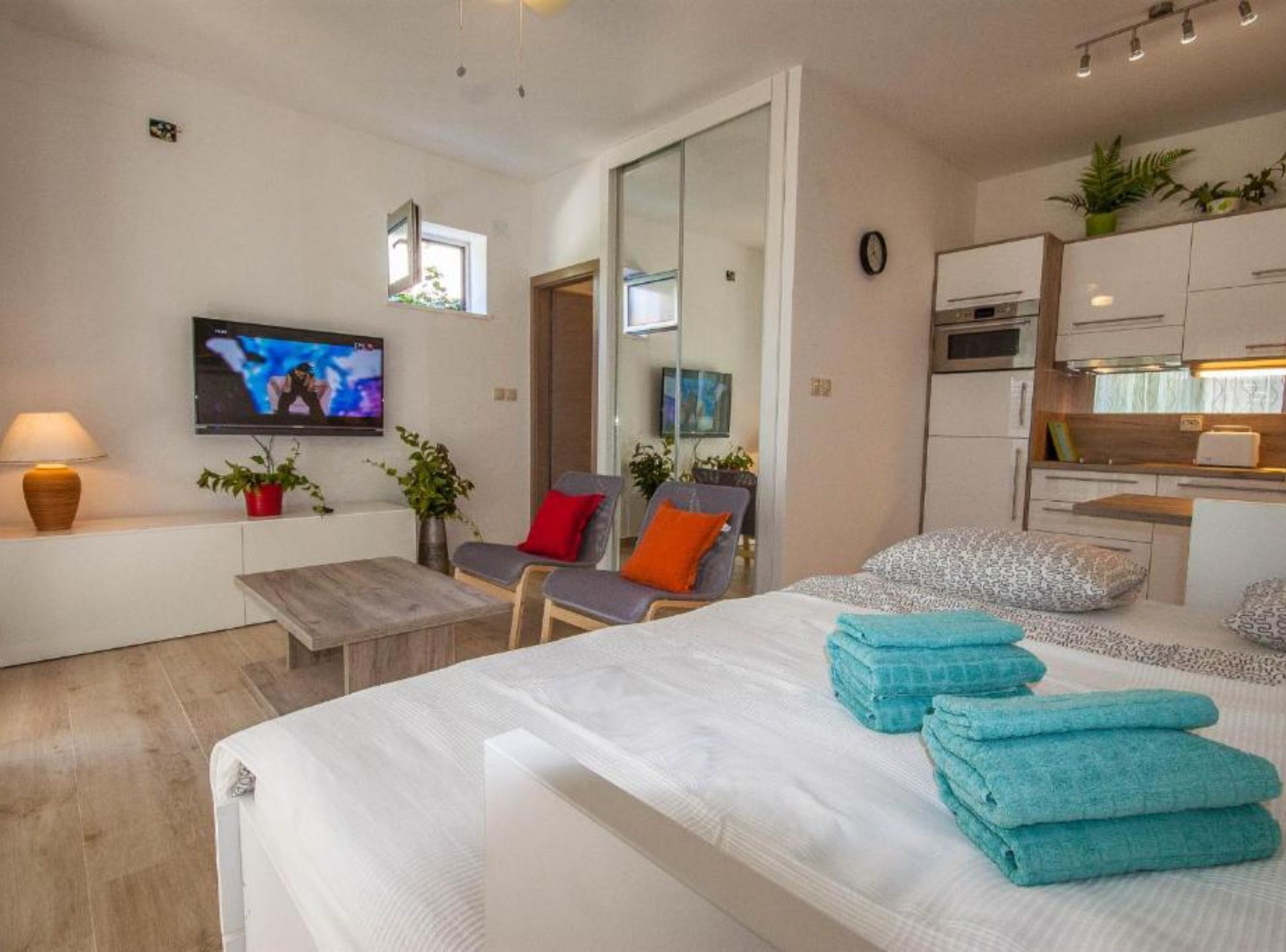 A modern hotel room with a bed, seating area, TV, kitchenette, and a large mirror.