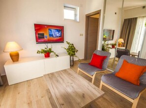Ein modernes Wohnzimmer mit einem Flachbildfernseher, zwei Sesseln mit roten Kissen, einem weißen Konsolentisch, einer Stehlampe und einem Topf mit Pflanze.