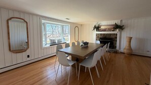 Uma sala de jantar com uma grande mesa de madeira, cadeiras brancas, uma lareira e um espelho grande na parede.