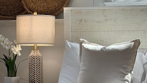 Uma cama bem arrumada com roupa de cama branca, uma luminária de cabeceira, uma planta em vaso e uma parede texturizada.
