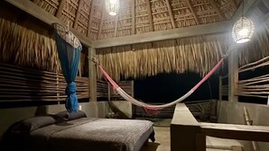 Ein Zimmer mit Strohdach, einer Hängematte, einem Bett und Blick auf den Nachthimmel.