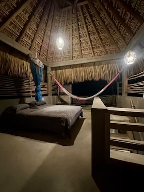 Una habitación con techo de paja, una hamaca, una cama y vistas al cielo nocturno.