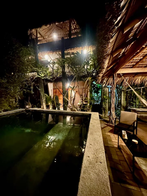Zona de piscina exterior con una estructura de techo de paja, rodeada de abundante vegetación.