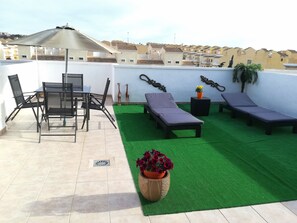 Eine Dachterrasse mit Esstisch, Stühlen, einer Liege, einem kleinen Tisch mit Topfpflanze und Kunstgras.