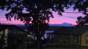 Uma casa com uma grande árvore em frente, um pequeno alpendre e vista para um corpo d'água e montanhas ao fundo.