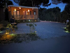 Een houten chalet met een veranda, een grindpad en omliggende begroeiing.