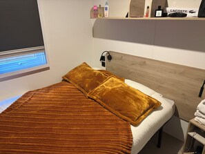 Een netjes opgemaakt bed met een bruin plaid en kussens, een nachtkastje met een lamp, en een plank met toiletartikelen en een Lacoste tas.