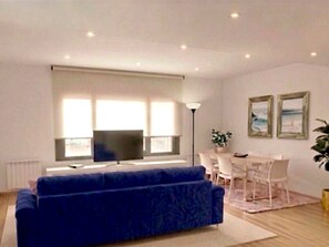 Ein modernes Wohnzimmer mit einer Couch, einem Fernseher, einem Esstisch, Stühlen und einem großen Fenster.