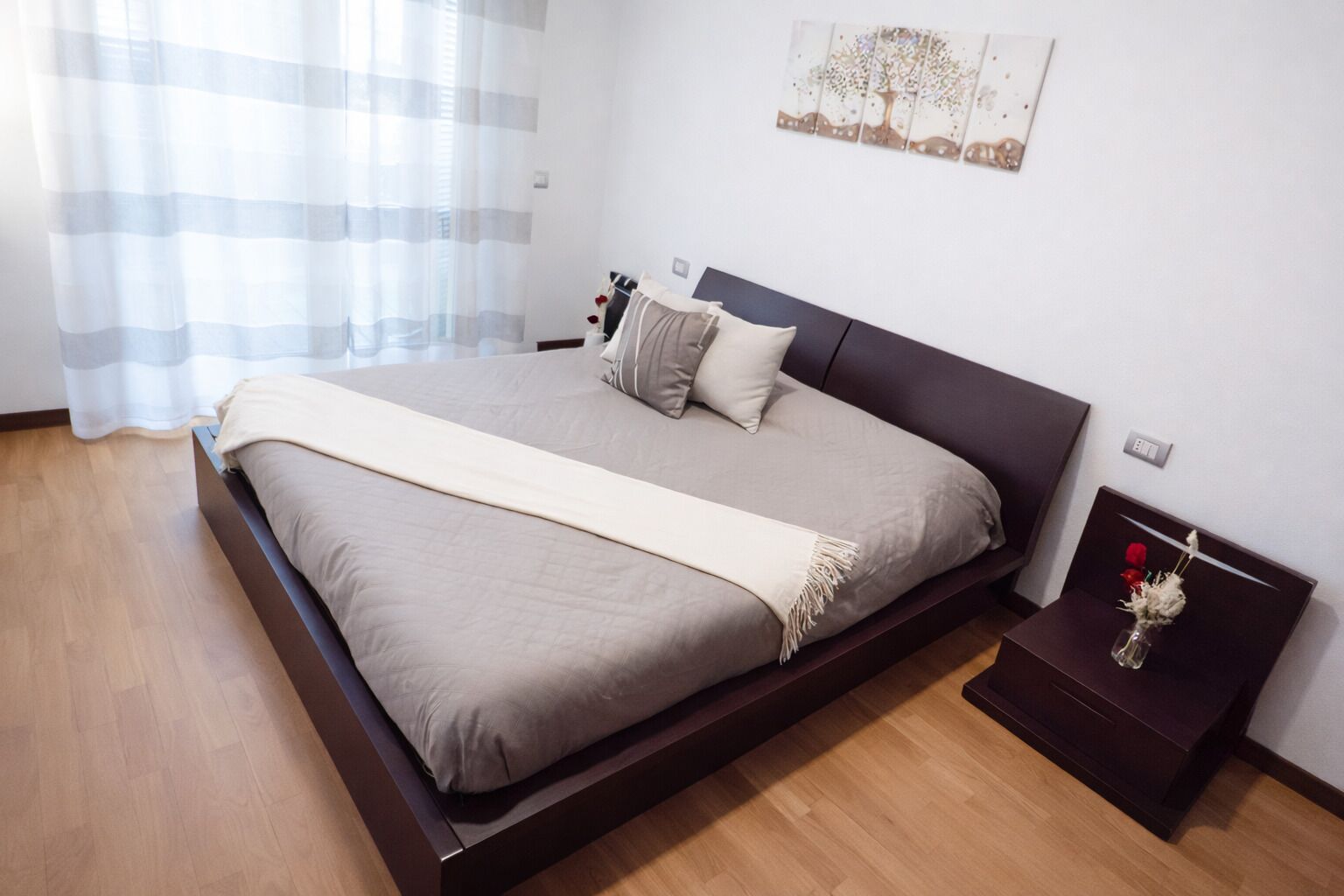 Un dormitorio con cama, mesita de noche, una obra de arte en la pared y ventana con cortinas.
