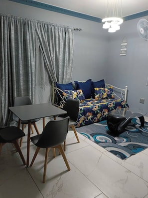 A room with a bed, a table with chairs, a fan, and a decorative rug.