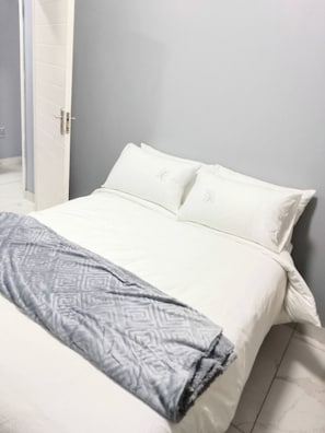 A bed with white bedding and a patterned throw, a grey wall, and a door leading to a bathroom. - Crompton Apartments 786 (Durban)