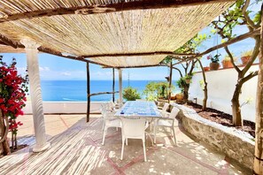 A covered outdoor seating area with a table and chairs, overlooking a body of water. - Caravella Nina Praiano  (PRAIANO)