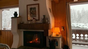 A cozy room with a fireplace, wooden beams, a window with a snowy view, and a lamp on a side table. - Our family chalet in the heart of the Alps (Chamonix-Mont-Blanc)