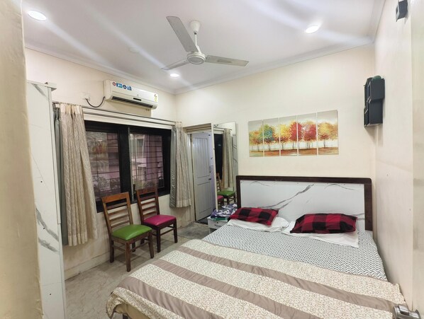 A bedroom with a bed, a ceiling fan, an air conditioner, a window with curtains, and a painting on the wall.