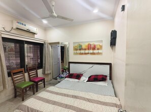 Ein Schlafzimmer mit einem Bett, einem Deckenventilator, einer Klimaanlage, einem Fenster mit Vorhängen, einem Sessel und einem Bild an der Wand.