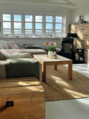 A cozy living room with a sofa, a wooden coffee table, a fireplace, and a view through large windows.