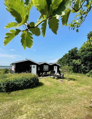 A black cabin with a patio, surrounded by greenery and a clear sky.