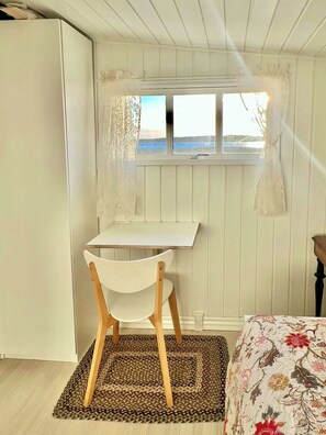 Une pièce avec un bureau en bois, une chaise blanche, un lit avec une literie à motifs floraux et une vue sur la mer par la fenêtre.