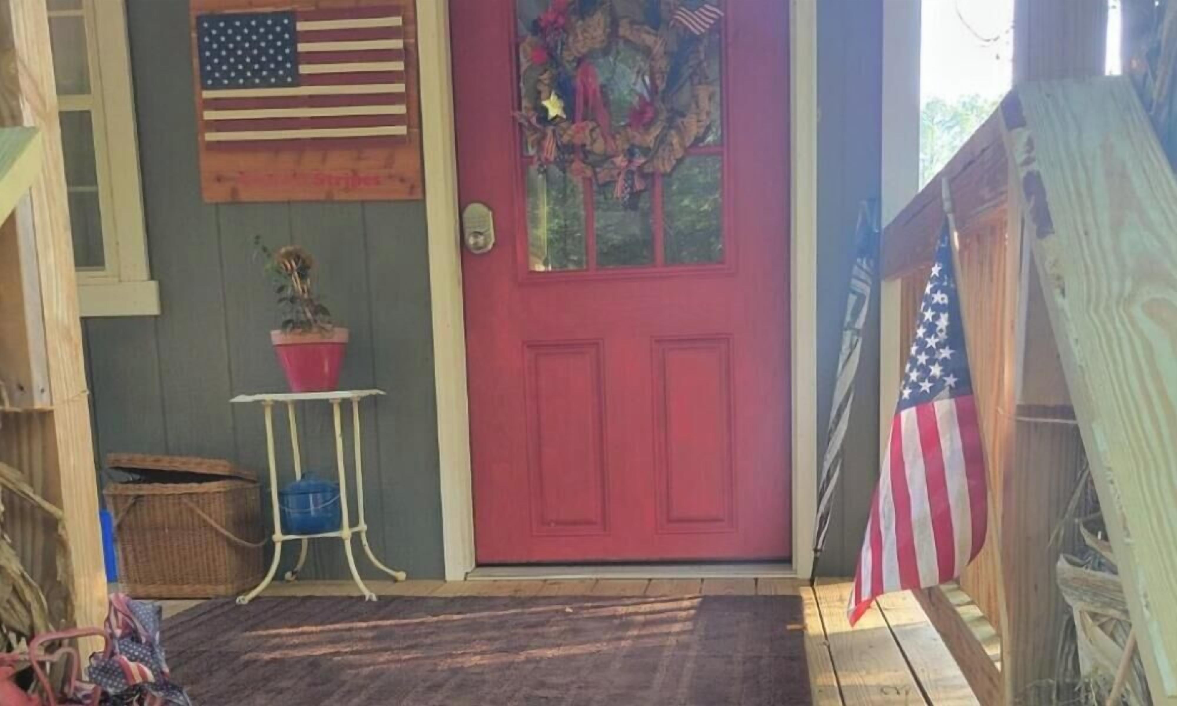 A red door with an American flag and a wreath, a small table with a potted plant, and a wooden fence.
