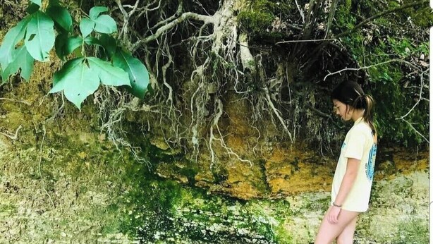 Eine Person steht an einem klaren Gewässer, umgeben von dichter Vegetation und felsigem Gelände.