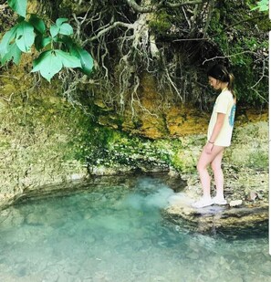 A person standing by a clear water body surrounded by natural vegetation and rocky terrain.