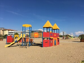 Une aire de jeux de plage avec des toboggans et des structures d’escalade, le tout sur fond de bâtiments et sous un ciel dégagé.