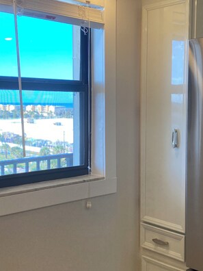 Eine Küche mit einem Edelstahl-Kühlschrank, einem Fenster mit Jalousien und Blick auf einen Parkplatz sowie umliegende Gebäude.