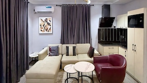 A modern living room with a sofa, a red armchair, a coffee table, a ceiling light fixture, and a kitchen area with cabinets and appliances. - Citiscape Apartment (Ikeja)