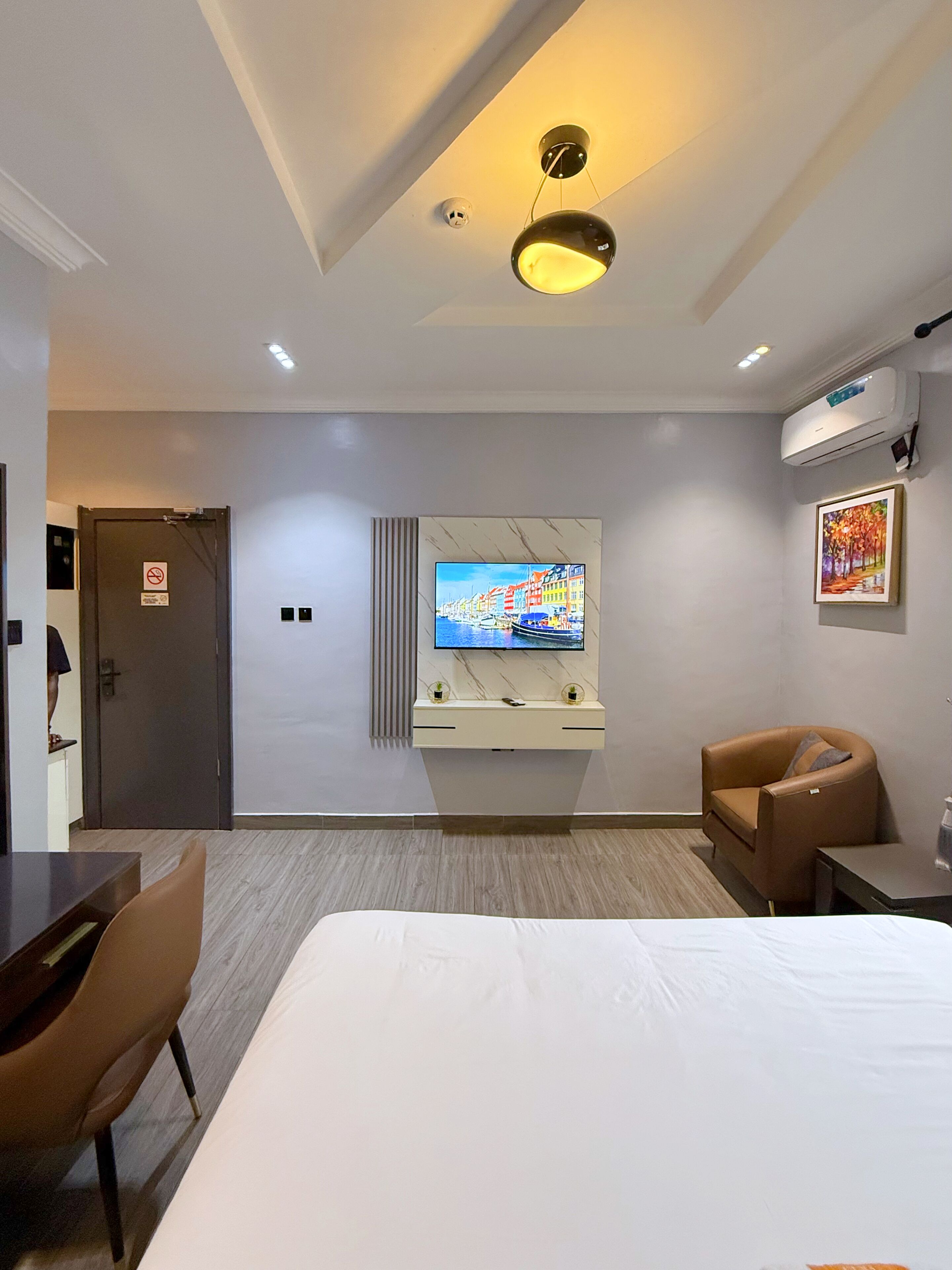 A hotel room with a bed, a TV mounted on the wall, a chair, a desk, and an air conditioning unit.