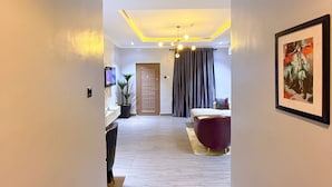 A hotel hallway with a wooden floor, a TV, a desk with a computer, a chair, a bed, a window with curtains, and a framed picture on the wall. - Citiscape Apartment (Ikeja)