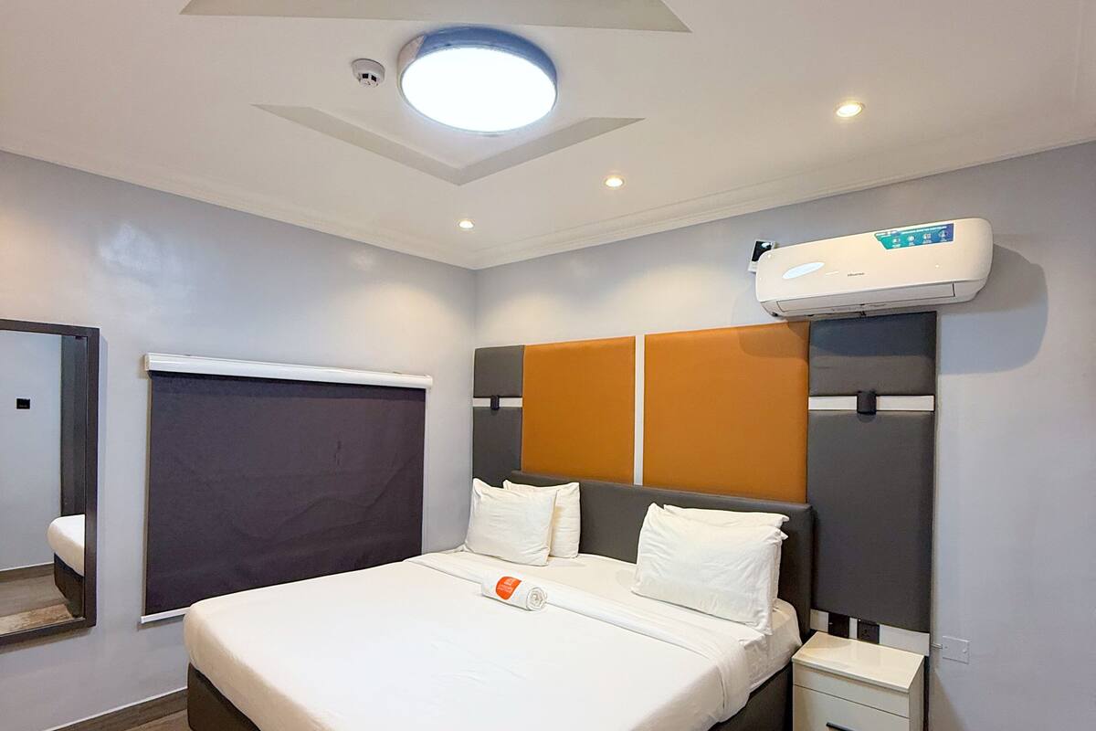 Una habitación de hotel moderna con una cama grande, una mesita de noche, un aire acondicionado de pared y una lámpara de techo.