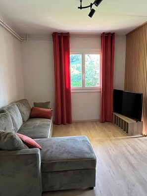 Een woonkamer met een bank, een poef, een televisie en een raam met gordijnen.