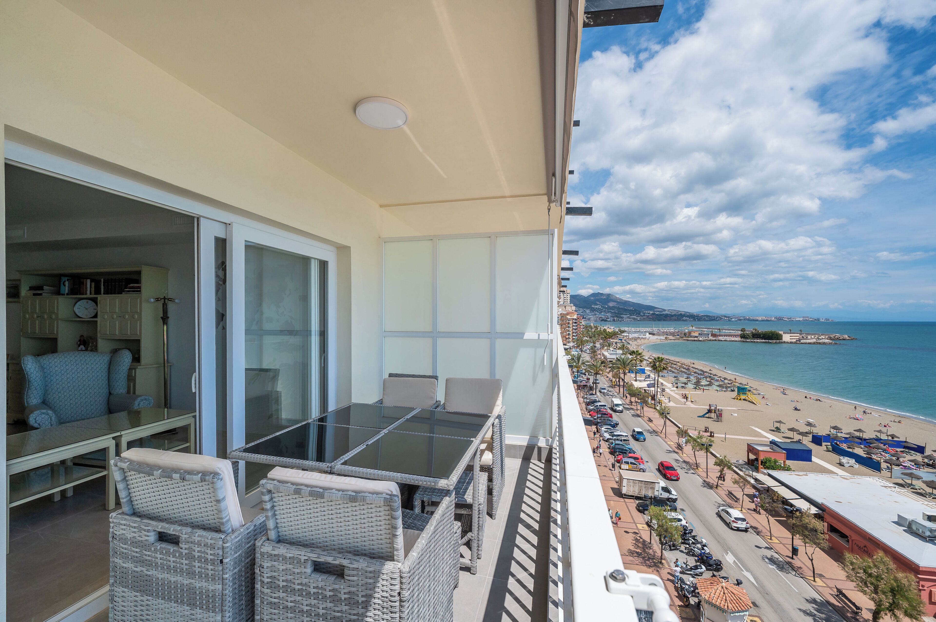 A balcony with a glass table and wicker chairs, overlooking a beach with parked cars and buildings.