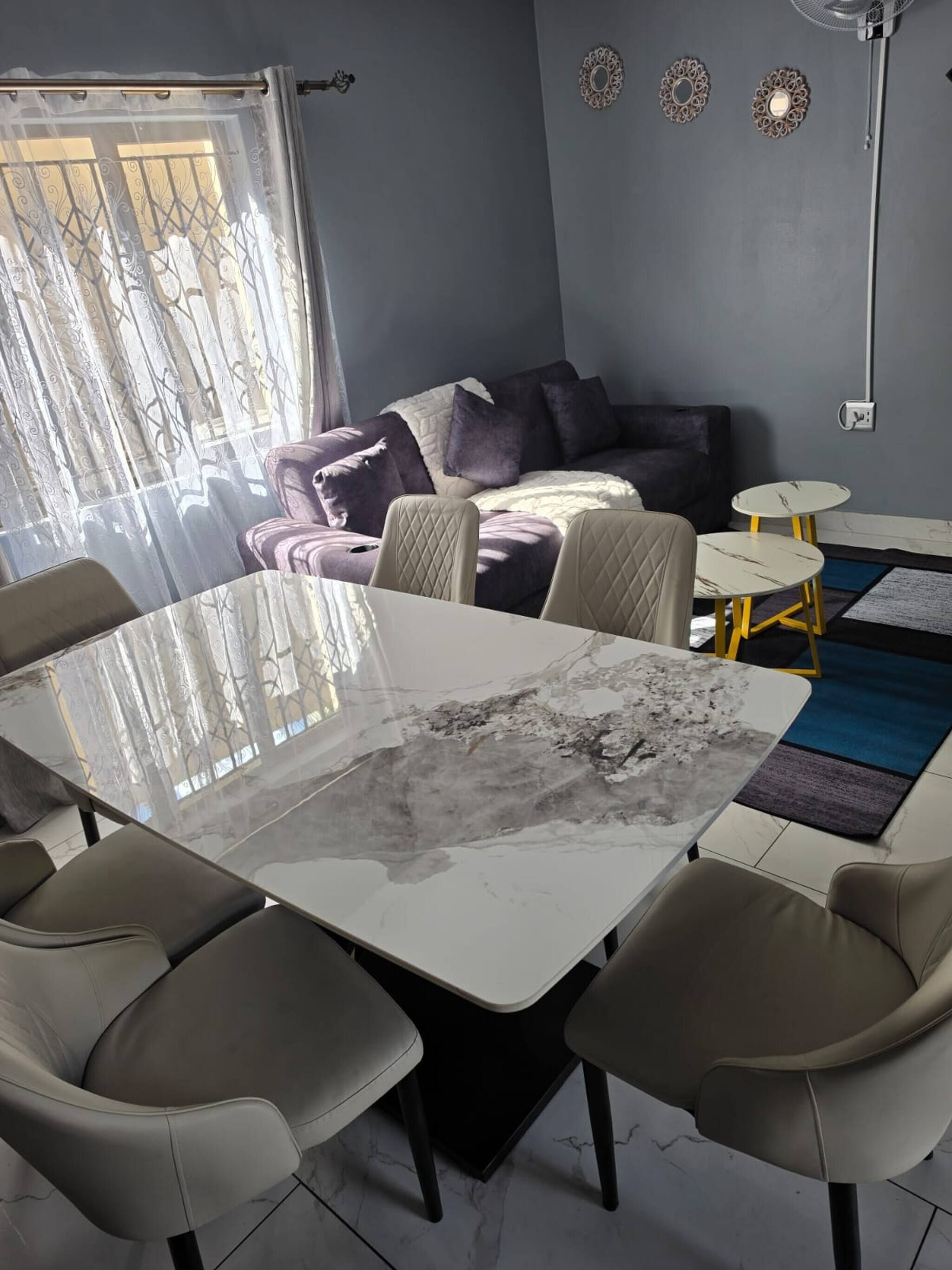 A modern living room with a glass-top dining table, beige chairs, a sofa, and a small coffee table.