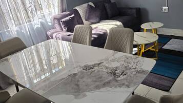 A modern living room with a glass-top dining table, beige chairs, a sofa, and a small coffee table.