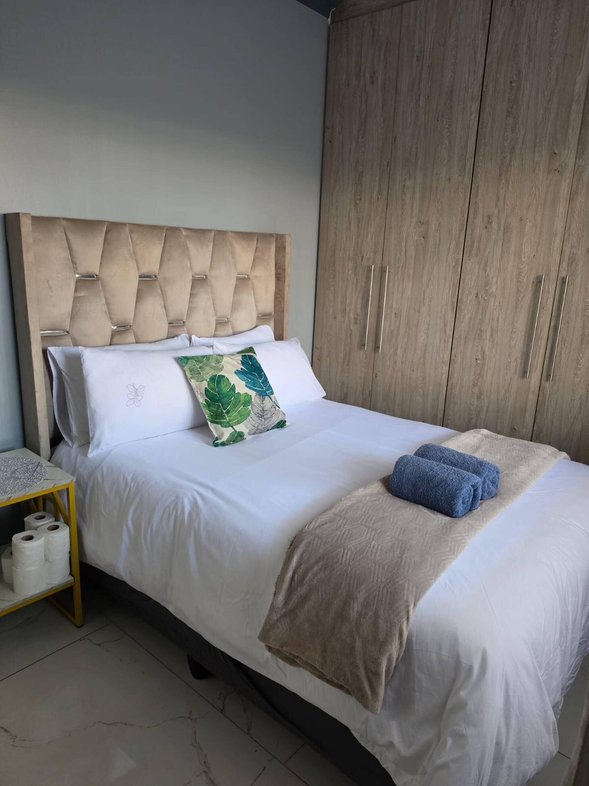 Un lit bien fait, doté d’une tête de lit capitonnée, une table de chevet avec un rouleau de papier toilette et une armoire à deux portes.