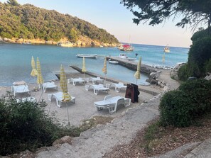 Ein Strand mit Liegestühlen und Sonnenschirmen, eine Anlegestelle mit Booten und ein bewaldeter Hügel.